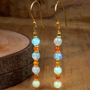 Pode incluir: Um par de brincos dourados com um design vertical. Cada brinco apresenta uma série de contas redondas de opala iridescente e contas laranja, enfiadas juntas. Os brincos têm um fecho de gancho.