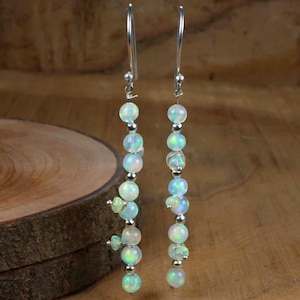 May include: A pair of long dangle earrings featuring iridescent opal beads and small silver beads. The earrings have a silver hook and are displayed against a wooden background. The opal beads have a mix of white, green, and blue hues.