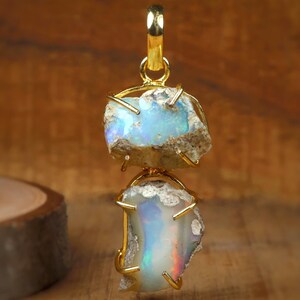 May include: A gold pendant featuring two rough-cut opal stones. The opals display iridescent flashes of blue, green, and red. The stones are held in place by a gold setting, with a gold loop at the top for hanging. The background is a warm, wooden surface.