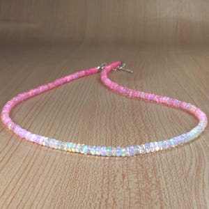 Puede incluir: Un delicado collar compuesto por pequeñas cuentas iridiscentes. El collar pasa de un tono rosa vibrante a un blanco opalescente brillante. Las cuentas están ensartadas, formando una curva elegante. El collar se muestra sobre una superficie de madera.