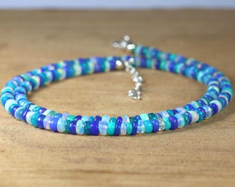 May include: A beaded necklace with a silver clasp. The necklace is made of small, round beads in shades of blue, teal, and white. The beads are strung together to form a double strand. The necklace is displayed on a wooden surface.
