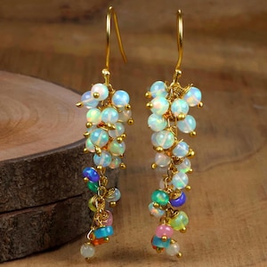 May include: Gold-tone drop earrings with clusters of iridescent opal beads and colourful gemstone accents. The earrings have a hook closure and are designed to be lightweight. These earrings are a unique statement piece, suitable for various occasions.
