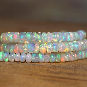 Puede incluir: Primer plano de tres hilos de cuentas de ópalo blanco. Las cuentas son redondas y tienen una sutil iridiscencia, mostrando destellos de color que incluyen verde, azul y naranja. Las cuentas están ensartadas, creando un collar o pulsera.