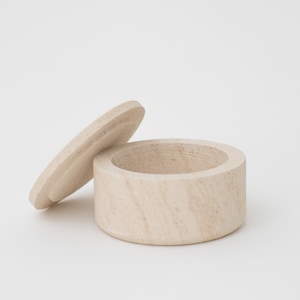 May include: A small, round, wooden box with a lid. The box is made of light-colored wood and has a natural finish. The lid is slightly ajar, revealing the empty interior of the box.