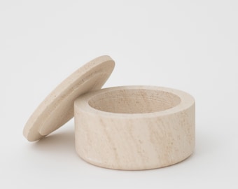 May include: A small, round, wooden box with a lid. The box is made of light-colored wood and has a natural finish. The lid is slightly ajar, revealing the empty interior of the box.