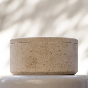May include: A round, beige, concrete container with a lid. The container has a rough, textured surface.