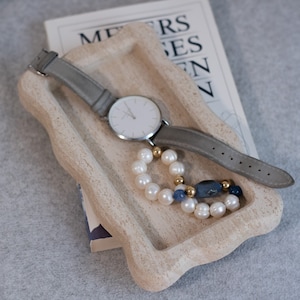 Peut inclure: Une montre gris clair avec un cadran blanc et un bracelet argenté se trouve dans un plateau beige avec un bracelet de perles et de perles bleues. Le plateau est fait d'un matériau texturé et a un bord ondulé.
