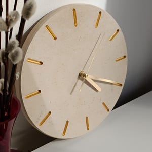 May include: A round, off-white wall clock with gold-colored hour markers and hands. The clock has a textured surface.