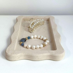 May include: A white stone tray with a wavy edge, featuring a gold hair clip with rhinestones and a pearl bracelet with blue accents.