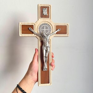 May include: A wooden crucifix featuring a silver-toned metal figure of Jesus Christ centred on the cross. The cross has a light-coloured border, dark wood panels, a circular Saint Benedict medal behind the figure, and an INRI scroll at the top.