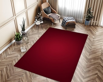 May include: A large, rectangular area rug featuring a solid, deep crimson red colour. The rug is positioned centrally on a light-toned wooden floor arranged in a herringbone pattern, within a room containing a chair, ottoman, and plants.