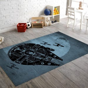 May include: A rectangular area rug featuring a large, black, detailed silhouette of a circular spaceship and two smaller fighter craft against a textured, circular, light blue background that resembles a moon or planet surface.