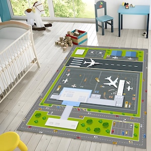 Puede incluir: Una alfombra de juego gris y verde con un diseño de aeropuerto detallado. La alfombra presenta una pista, calles de rodaje, un edificio terminal y aviones. También hay coches y camiones pequeños en la alfombra.