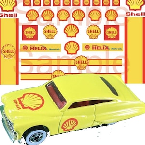 May include: A yellow toy car with Shell logos and decals. The car has a red interior and black wheels. The car is a 1950s style coupe.