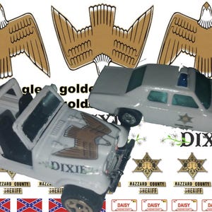 May include: Two toy cars, a white Jeep and a white police car, with a golden eagle design on the Jeep. The cars are surrounded by several gold stars with the text "Hazzard County Sheriff" and "Dixie" printed on them.