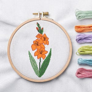 May include: Cross-stitch embroidery of an orange gladiolus flower with green leaves, set in a wooden hoop. The needlework is on white fabric. Colorful embroidery floss in shades of green, orange, purple, yellow, blue, and pink are visible.