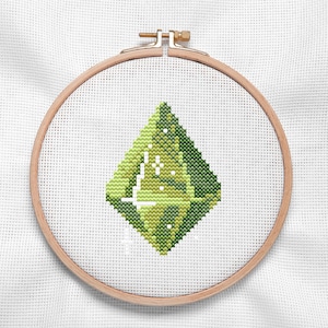 May include: A cross-stitch embroidery project featuring a diamond-shaped gemstone design in shades of lime, olive, and forest green. The pattern is centered on white Aida fabric and secured within a round wooden embroidery hoop with a metal screw.