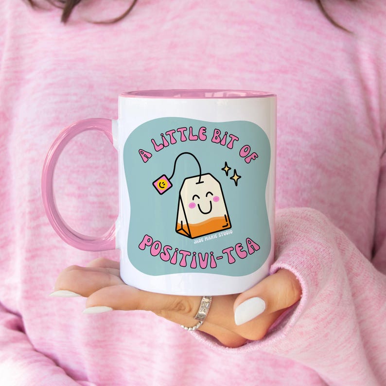 May include: White ceramic mug with a pink handle and interior. The mug features the text "A LITTLE BIT OF POSITIVI-TEA" with a smiling tea bag graphic on a light blue background. The mug is held by a person wearing a pink sweater.