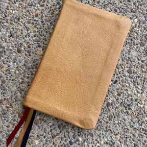 May include: A tan leather book cover with a textured surface. Three colored ribbons, red, tan, and navy, extend from the bottom. The cover is rectangular with rounded corners.