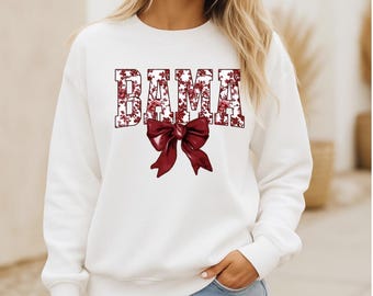 May include: White sweatshirt with the word "BAMA" in a floral design and a burgundy bow graphic. The sweatshirt is a crew neck style and is made of a soft material.