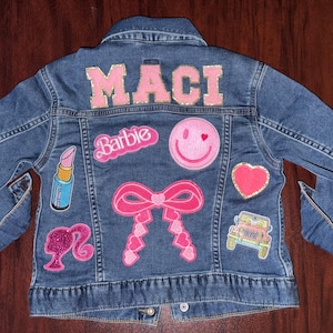 May include: A blue denim jacket with the name "MACI" in pink letters across the back. The jacket is decorated with pink patches including a smiley face, a heart, a bow, and the word "Barbie".