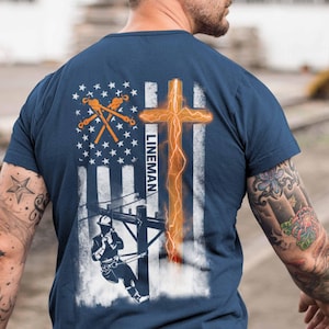 May include: Navy blue t-shirt featuring a graphic design of an American flag with a cross and a lineman at work. The design includes the word "LINEMAN" and utility tools. The shirt is made of a soft material.