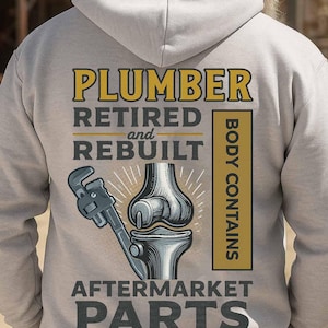 May include: Light gray hoodie with the text "PLUMBER RETIRED and REBUILT" in yellow and black. A graphic of a wrench and a knee joint is below the text. The phrase "BODY CONTAINS AFTERMARKET PARTS" is also printed on the back.