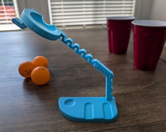 May include: A blue plastic pong ball launcher with a base and a long arm that extends up and over. The arm has the word "PONGER" printed on it. There are three orange ping pong balls in front of the launcher and two red plastic cups behind it.
