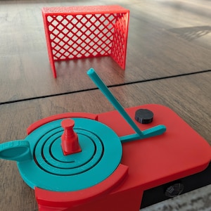 Puede incluir: Un juego de hockey de mesa rojo y turquesa. El juego incluye una portería roja, una base de juego turquesa y roja con un asa turquesa, un palo de hockey turquesa, un disco rojo y una pieza central roja. El juego es de plástico.