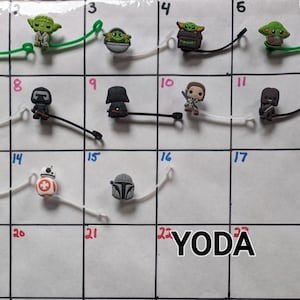 Puede incluir: Una cuadrícula blanca con los números del 1 al 24, cada cuadrado contiene un amuleto de personaje diferente de Star Wars. Los amuletos están hechos de goma y presentan personajes como Yoda, Darth Vader y R2-D2.