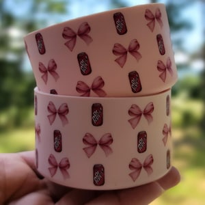 May include: Two stacked, light pink containers with a repeating pattern of maroon bows and Dr. Pepper cans. The containers are cylindrical and appear to be made of a smooth material. The background is a blurred view of green foliage.