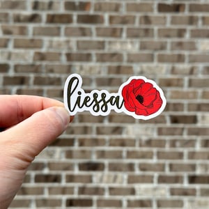 May include: A sticker with the word "liessa" written in cursive in brown and a red poppy flower.