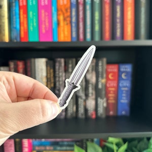 May include: A sticker of a silver dagger with a detailed handle. The sticker is held in front of a bookshelf filled with colorful books. The dagger has a pointed blade and ornate design on the handle.