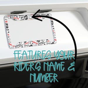 Peut inclure: Cadre de plaque d'immatriculation de moto blanc avec une bordure noire et un motif blanc et noir de graphismes sur le th&egrave;me de la moto. Le cadre pr&eacute;sente un espace clair pour le nom et le num&eacute;ro du pilote. Le texte "FEATURES YOUR RIDER'S NAME & NUMBER" est &eacute;crit en bleu turquoise.