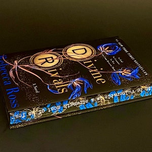 Può includere: Un libro rilegato in brossura dal titolo "Divine Rivals" di Rebecca Ross. La copertina presenta uno sfondo nero con disegni floreali dorati e blu. Il libro è un best-seller del New York Times.