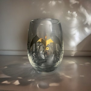 Op de afbeelding: Een helder glazen wijnglas met een gouden drakenontwerp. De draak vliegt met zijn vleugels wijd uitgespreid. Het glas staat op een wit oppervlak.