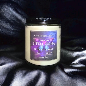 Può includere: Candela in cera di soia in un barattolo di vetro con coperchio nero. L'etichetta presenta il testo "smells like LITTLE OSHA" con un disegno a farfalla. La candela pesa 227 g e ha una durata di combustione di oltre 40 ore. Sfondo scuro in tessuto serico.