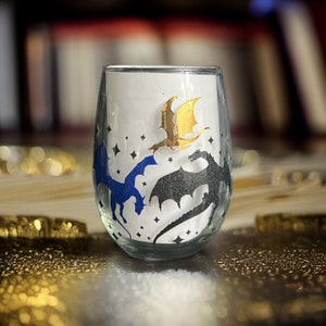 Op de afbeelding: Een helder glazen wijnglas met een ontwerp van drie draken. De draken zijn in tinten blauw, zwart en goud. Het glas is perfect om van uw favoriete drankje te genieten.