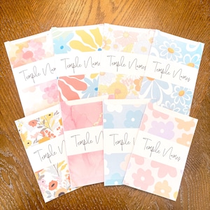 Op de afbeelding: Een verzameling notebooks met bloemmotieven en de tekst "Temple Names". De ontwerpen omvatten madeliefjes, abstracte bloemen en aquareleffecten in pasteltinten. Gerangschikt in een waaier.