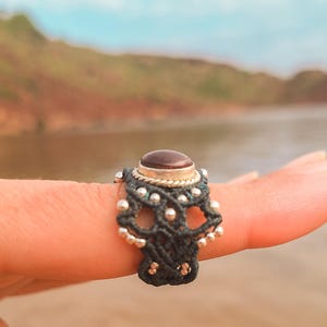 Pode incluir: Um anel artesanal com uma pedra preciosa escura em uma banda de prata e ouro. O anel tem uma banda em estilo macramê na cor verde-azulado escuro, com contas de prata adicionando detalhes. O anel é mostrado em um dedo.