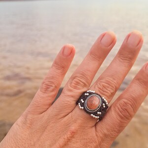 Peut inclure: Bague artisanale ornée d'une pierre précieuse ovale de couleur pêche, sertie dans une bande en macramé marron foncé. La bande est ornée de petites perles argentées. La bague est présentée sur une main, avec un arrière-plan flou d'eau et de ciel.