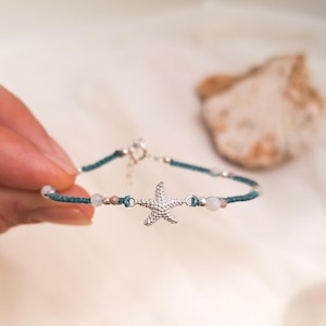 Può includere: Un braccialetto in corda blu turchese con un ciondolo a forma di stella marina argentata e perline bianche e rosa.