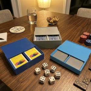 Könnte beinhalten: Drei rechteckige Aufbewahrungsboxen mit Spielkarten, bestehend aus einer dunkelblauen, einer hellblauen und einer grauen Box. Sechs weiße sechsseitige Würfel mit schwarzen Punkten sind vor den Boxen auf einer Holzoberfläche angeordnet.