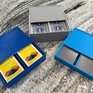 Könnte beinhalten: Drei rechteckige Kartenaufbewahrungsboxen aus Kunststoff mit Schiebedeckeln. Eine graue Box enthält blaue Spielkarten. Eine dunkelblaue Box enthält gelbe Karten mit dem Text "TO THE TOP". Eine hellblaue Box enthält graue Karten mit dem Text "of 3's".