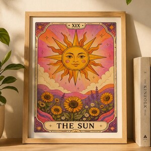 May include: A framed tarot card print titled THE SUN with the Roman numeral XIX. The illustration features a large yellow sun with a face, radiating light over a field of sunflowers and purple flowers against a pink and orange sky.