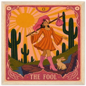 May include: A framed tarot card print titled THE FOOL featuring a person in an orange striped dress and pink boots walking through a desert with cacti, a bright sun, and a small rabbit. The person carries a stick with a pink bundle over one shoulder.