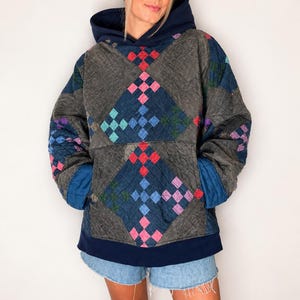 Puede incluir: Una sudadera con capucha y manga larga con un diseño de patchwork acolchado. La prenda presenta una base gris oscuro con patrones en forma de diamante en rojo, azul, rosa y verde. Incluye una capucha, puños y dobladillo azul marino.