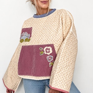 Pode incluir: Um suéter acolchoado de manga comprida em tecido cor de creme com um pequeno padrão floral. Inclui um bolso frontal grande em ganga malva escuro e um bolso de peito mais pequeno a condizer, ambos decorados com aplicações de flores e folhas.