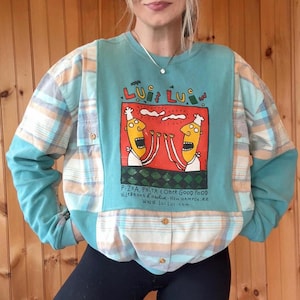 May include: A teal and plaid patchwork sweatshirt with a graphic of two cartoon chefs. The graphic is in a red square and the text reads "Lui Lui" and "Pizza, Pasta & Other Good Food". The sleeves are plaid.