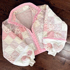 Può includere: Una giacca trapuntata con un design patchwork nei toni del rosa, bianco e motivi floreali. La giacca ha un bordo rosa e fiocchi sulle maniche. L'indumento è appoggiato su una superficie di legno scuro.